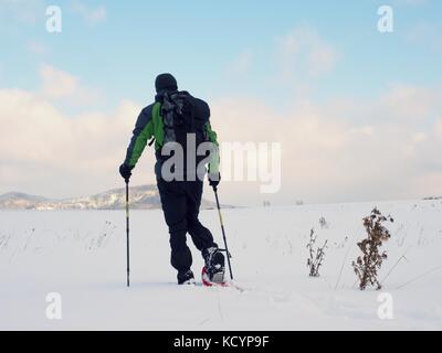 Mann mit Schneeschuhen Spaziergang in verschneiter eingereicht. Wanderer in Grün Grau winter Jacke und Hose schwarz trekking Schneeschuhwandern im Pulverschnee. bewölkt Wintertag, g Stockfoto