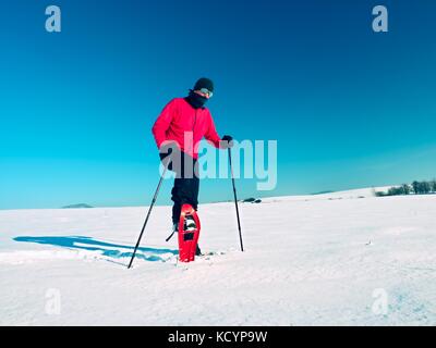 Mann Reinigung Snowshoe. winter Tourist mit Schneeschuhen gehen in Snowy Drift. sonnigen freeze Wetter. Wanderer in rosa Sakko und Hose schwarz Trekking Stockfoto