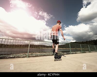 Mittleres Alter Mann im roten T-Shirt mit Inline Skates fahren im Sommer Park, populäre outdoor Skaten. Meer Brücke mit blauem Himmel in backgrround Stockfoto