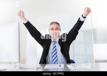 Gerne reife Geschäftsmann Anheben der Arme auf dem Schreibtisch im Büro Stockfoto