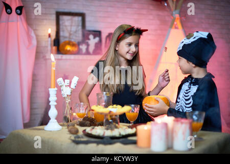Porträt von zwei Kinder, Junge und Mädchen tragen Halloween Kostüme plaudern während der Partei stehend am Tisch mit Süßigkeiten und Snacks in eingerichtete Zimmer Stockfoto