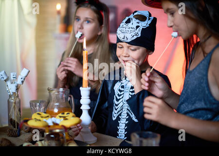 Portrait von Stattlichen kleinen Jungen tragen Skelett Kostüm genießen Lecker gebratenen Marshmallows während Halloween Feiern mit Freundinnen, Innenraum o Stockfoto