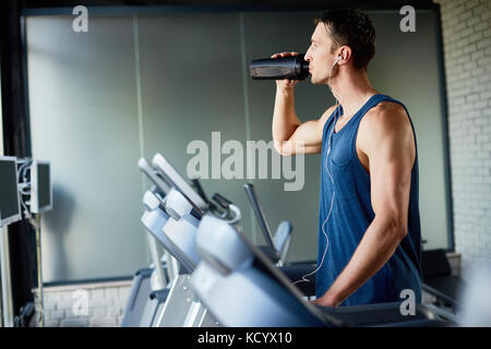 Konzentriert Sportsman in Kopfhörer selbst erfrischend mit Wasser während der Ausführung auf Laufband im Fitnessstudio, Profil anzeigen Stockfoto