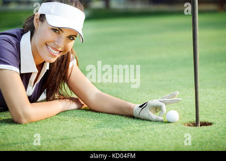 Glückliche junge Frau drückt Golf Golf Ball in das Loch, liegend auf grünem Gras Stockfoto
