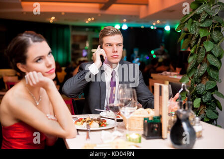 Junge Frau gelangweilt auf dem neuesten Stand, während ihr Freund in der Zelle auf dem Mobiltelefon Stockfoto