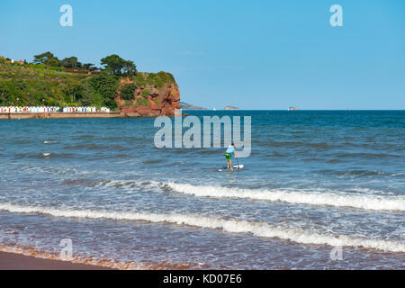 Paddel - boarder Position heraus zum Meer bei Goodrington Sands, Paignton, Devon. de.Strandhütten und roten Klippen am roundham Kopf mit Boot in der Ferne Stockfoto