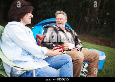 Reifes Paar sitzen in camping Stühle, neben Zelt, holding Tassen Tee, lächelnd Stockfoto