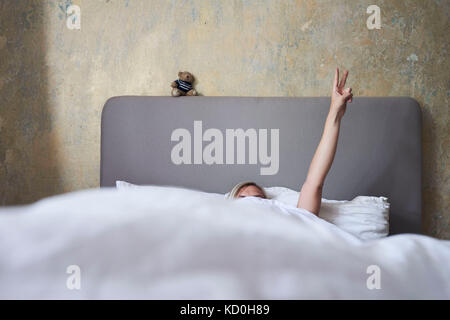 Frau im Bett, versteckt sich unter Abdeckungen, Arm in der Luft, Hand, Frieden Zeichen Stockfoto