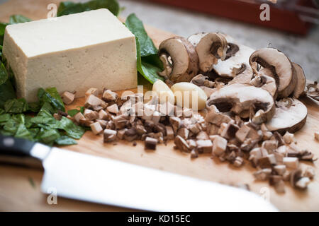 Gehackte und geschnittene Pilze, Spinat und Tofu auf einem Schneidebrett mit einem Messer. Stockfoto