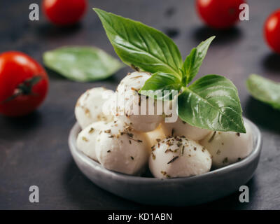 Nahaufnahme der kleinen Mozzarella Käse mit Kräutern mariniert. Mozarella Kugeln mit frischem Basilikum und Tomaten auf Schwarz konkreten Hintergrund. Niedrig Stockfoto