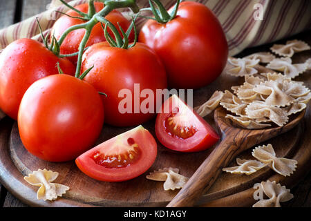 Frische rote reife Tomaten auf der Rebe auf einem dunklen rustikalen Schneidbrett mit Vollkorn Bow Tie Pasta Stockfoto