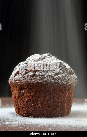 Nahaufnahme eines köstlichen Schokolade muffin auf einem Holztisch, auf schwarzem Hintergrund, mit Schnee wie Pulver Zucker um und über fallende verstreut. Stockfoto