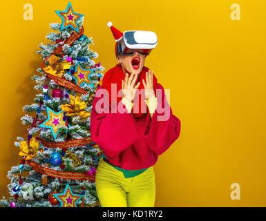 Festliche Jahreszeit. überrascht, moderne Frau in bunten Kleidern in der Nähe von Weihnachten Baum auf gelben Hintergrund tragen Virtuelle Realität Gläser Stockfoto