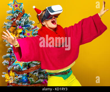 Festliche Jahreszeit. Freundliche junge Frau in bunten Kleidern in der Nähe von Weihnachten Baum auf gelben Hintergrund tragen Virtuelle Realität Gläser Stockfoto