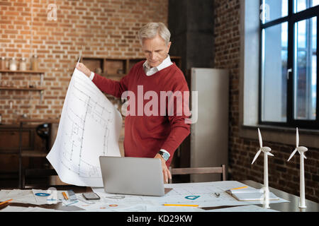 Grauhaariger Mann mit Blueprint und Tippen auf Laptop Stockfoto