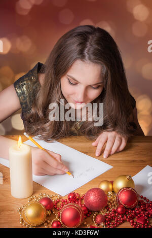 Junge schöne Mädchen schreiben zu Santa Claus, bei Kerzenlicht, mit bunten Kugeln und andere Weihnachtsdekorationen umgeben. Stockfoto