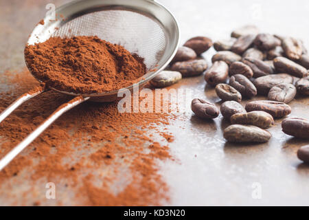 Dunkles Kakaopulver mit einem Sieb und Kakaobohnen auf alten Küchentisch. Stockfoto