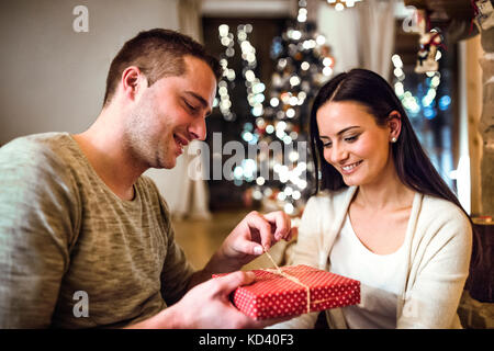 Schöne junge Paare sitzen auf dem Boden vor dem Kamin. Mann und Frau Geschenke geben. Stockfoto