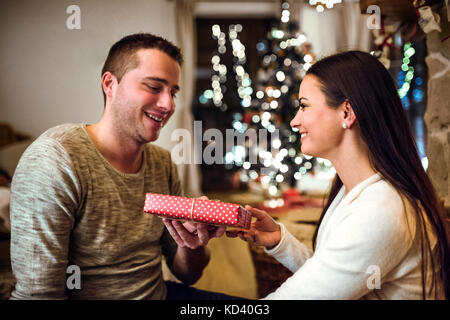 Schöne junge Paare sitzen auf dem Boden vor dem Kamin. Mann und Frau Geschenke geben. Stockfoto
