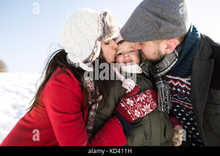 Vater und Mutter Spaß mit ihrer Tochter, die im Schnee spielen. sonnigen weißen Winter Natur. Eltern küssen ihre kleine Tochter. Stockfoto