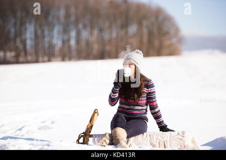 Schöne junge Frau auf gestrickte Mütze und Handschuhe mit einer Tasse Kaffee im Winter draußen Natur. Frau auf einem Schlitten sitzen. Stockfoto