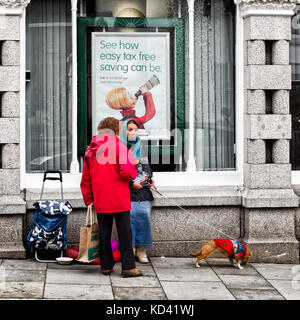 Eine junge Frau mit Kopftuch verkaufen das große Thema von einer Bank in Totnes Devon, mit einem Hund an der Leine, ein Einkaufswagen und ein Gespräch mit einem Kunden Stockfoto