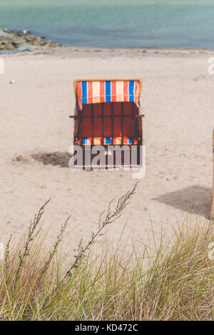 Leeres rotes überdachte Stuhl auf Sandstrand in Travemünde, Deutschland Nord. Trockenes Gras im Vordergrund Stockfoto