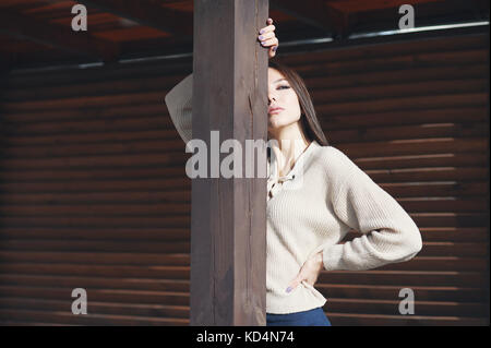 Porträt einer charmante junge Frau in einem Gewirke aus Wolle beige Pullover, auf einer hölzernen Hintergrund ausgehen. Blendet die Hälfte des Gesichts, und Posen vor der Kamera. Stockfoto