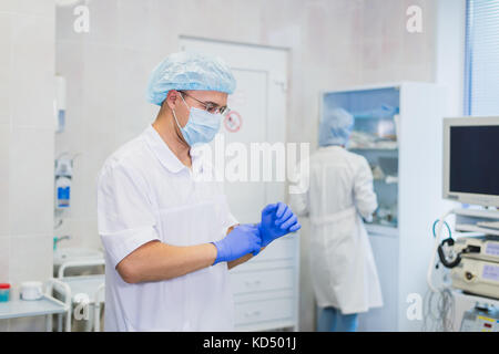 Junger Arzt während der Vorbereitung für den Betrieb. Assistentin der Chirurg das Tragen von Mundschutz in Betrieb Zimmer im Krankenhaus. Stockfoto