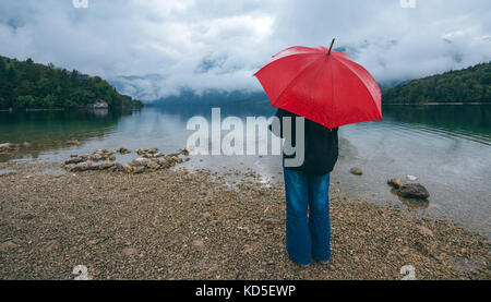 Frau mit roten Dach betrachtet auf Regen vor einem See. traurig und einsam weibliche Person in die Ferne schauen. Stockfoto