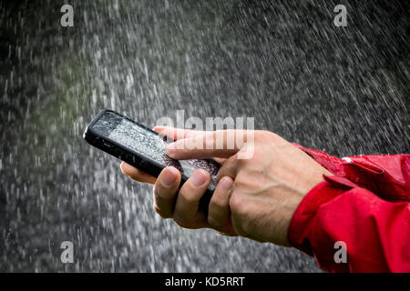 Ein Mann mit einem Mobiltelefon in einem wasserdichten Fall draußen im Regen Stockfoto