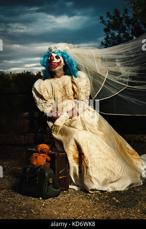 Ein unheimlich bösen Clown tragen eine schmutzige und ausgefranster Braut Kleid in einem Beunruhigenden ländliche Landschaft in der Dämmerung sitzen, neben ihr Gepäck und ein Teddybär Stockfoto