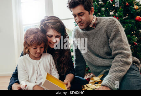 Kleine Familie sitzt in der Nähe von Weihnachten Baum unboxing Geschenke. Kleine Mädchen an ihr Weihnachtsgeschenk suchen. Stockfoto