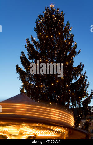 Ein sich drehendes Karussell unter dem Weihnachtsbaum auf Weihnachtsmarkt am Abend. Dekorative Xmas Tree mit Beleuchtung Merry-go-round bei Sonnenuntergang. Stockfoto