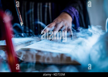 Nahaufnahme der Hand eine Hexe mit Spitzen schwarz Nägel auf smoky Buch der Magie Rezept und mit einem Zauberstab. Stockfoto