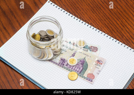 Glas voller Münzen auf Banknoten mit Notebook, auf hölzernen Schreibtisch Stockfoto