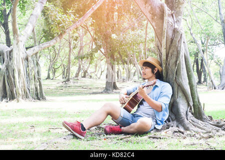 Happy asiatischer Mann sitzen unter Baum, tragen, Stroh, Hut und Gitarre spielen Stockfoto