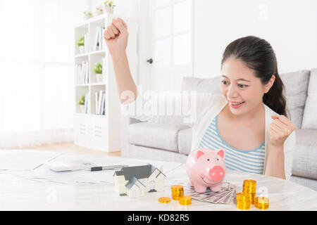 Glücklich lächelnde Frau speichern genug für die neue Wohnung kaufen können und das Sitzen im Wohnzimmer im Haus Modellbau erfolgreiche Geste Feier suchen. Stockfoto