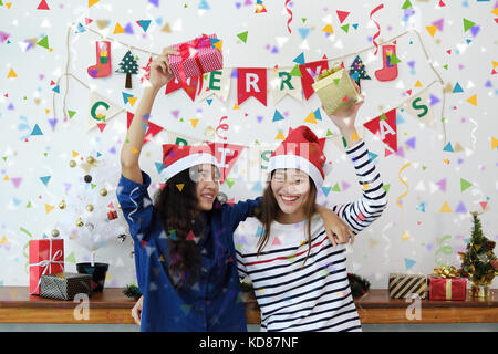 Zwei schöne junge Frauen in santa Hüte und Holding Geschenkboxen Spaß mit bunten Konfetti an der Weihnachtsfeier. Stockfoto