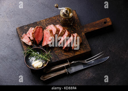 In Scheiben geschnitten Filet Steak Roastbeef mit Messer und Gabel Carving auf Holz Schneidebrett auf dunklem Hintergrund einstellen Stockfoto