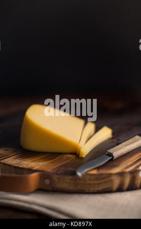 Keil aus geräuchertem gouda-Käse auf rustikalem Holzschneidebrett mit antikem Käsemesser aus Holz geschnitten. Stockfoto