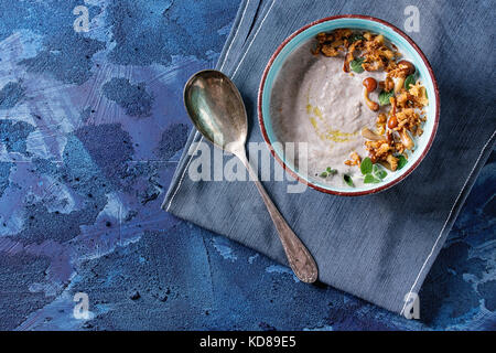 Pilz Cremesuppe in Keramik Schüssel mit Waldpilzen, grünen serviert, gebratenen Zwiebeln, Löffel auf Blau textil Serviette über Dunkelblau Textur konkrete Ba Stockfoto