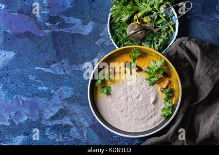 Pilz Cremesuppe in Schwarz Keramik Schüssel mit kurkuma Pulver, Waldpilzen, Grüns auf textile Serviette über Dunkelblau Textur konkrete zurück Stockfoto