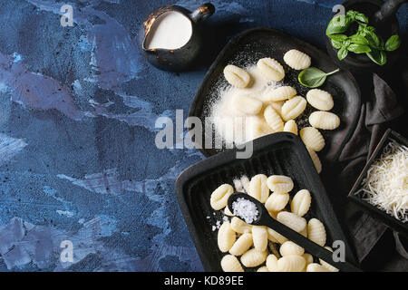 Rohen ungekochten Kartoffel Gnocchi in schwarz Holz- platten mit Zutaten. Mehl, geriebenen Parmesan, Basilikum, Salz, Krug von Creme über Dunkelblau Beton Stockfoto