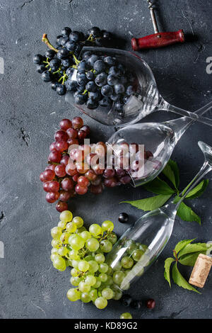 Vielzahl von drei Art frische reife Trauben dunkel Blau, Rot und Grün in verschiedenen Lügen Weingläser mit alten Korkenzieher über schwarzen Textur Hintergrund. Top Stockfoto