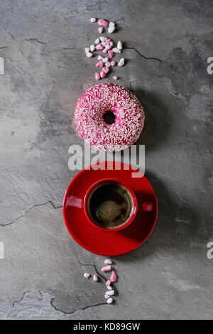 Rosa glasierten Krapfen mit rosa Zucker und rot Tasse schwarzen Kaffee über grau Textur Hintergrund. Flach mit Platz Stockfoto