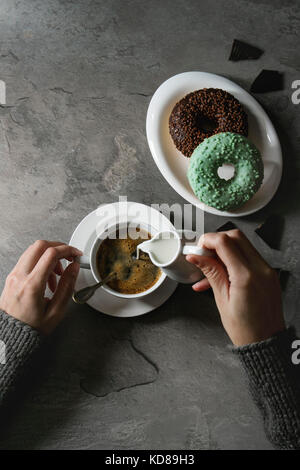 Platte von Bunt glasierter Krapfen mit gehackte Schokolade, Tasse schwarzen Kaffee, Kanne Milch über grau Textur Tabelle. Weibliche Hände goß Sahne. Flach Stockfoto