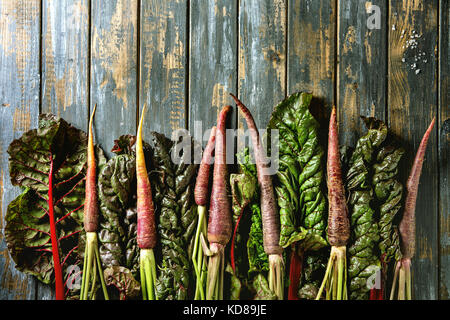 Raw organic lila Karotte mit Mangold mangold Blätter über alte hölzerne Planke Hintergrund. Ansicht von oben mit der Kopie. Essen Hintergrund. Stockfoto