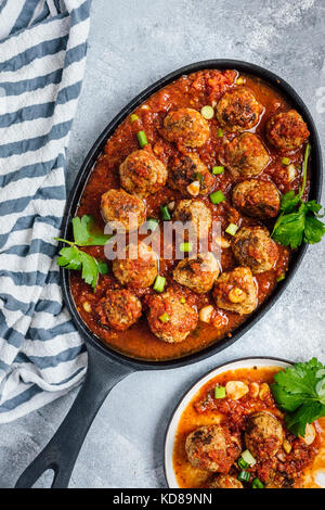 Rindfleisch Frikadellen in Tomatensoße in einer gusseisernen Pfanne gekocht auf grauem Hintergrund aus Sicht von oben fotografiert. Eine Platte mit Fleischbällchen begleitet. Stockfoto