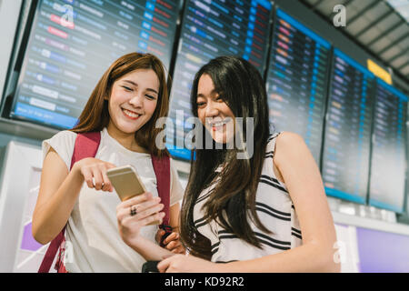 Zwei asiatische Mädchen mit zusammen an Flight Information Board im Flughafen smartphone. online Check-in, Fahrplan Anwendung oder Holiday Travel Concept Stockfoto